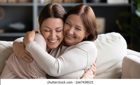 Affectionate loving middle aged mother cuddling grownup child, amazed by good news. Excited to see daughter, happy senior older mom cuddling her, sitting together on comfortable sofa in living room.