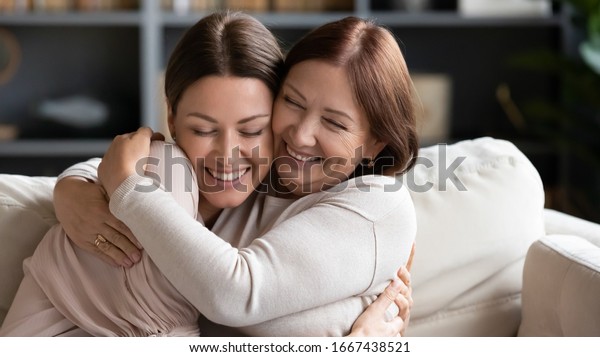 Affectionate loving middle aged mother cuddling\
grownup child, amazed by good news. Excited to see daughter, happy\
senior older mom cuddling her, sitting together on comfortable sofa\
in living room.