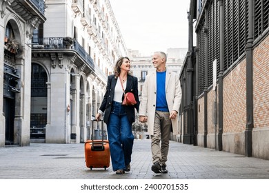 Affectionate mid adult tourist couple walking together with luggage in a european city. Lovely happy senior man and woman traveling with a suitcase visiting streets of old town. - Powered by Shutterstock