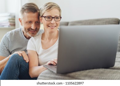 Affectionate Middle-aged Couple Looking At A Laptop Together On A Sofa In Their Living Room Browsing The Internet Or Social Media With A Smile