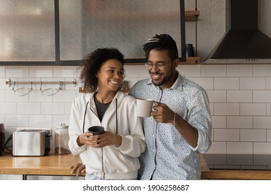 Affectionate Millennial African American Couple Meet Morning At Cozy Kitchen Embracing Talking Enjoying Hot Drinks. Happy Young Black Husband Wife Bonding Drinking Coffee Tea For Pleasant Conversation