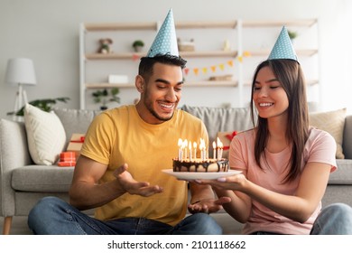 Affectionate millennial multiracial couple in party hats holding birthday cake with lit candles in living room. Positive diverse husband and wife celebrating b-day, spending holiday together