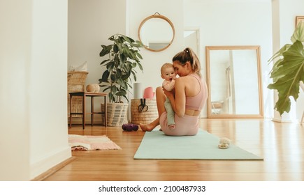 Affectionate mom working out with her baby at home. Healthy mom holding her baby while sitting on an exercise mat. New mom bonding with her baby during her post-natal fitness routine. - Shutterstock ID 2100487933