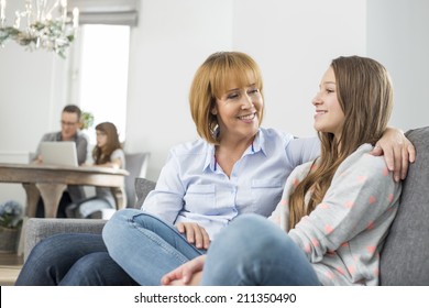 affectionate mother and daughter sitting on sofa with family in background