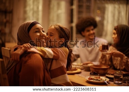 Affectionate Muslim grandmother and granddaughter embracing during family meal in dining room.