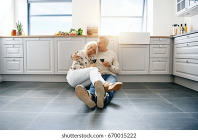 Affectionate senior couple smiling and sitting contently together on the floor of their bright kitchen at home drinking red wine - Powered by Shutterstock