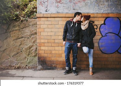 Affectionate Teenage Couple Kissing Outdoors Against A Wall On Street. Mixed Race Couple In Love.