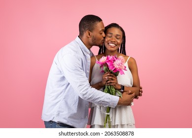 Affectionate young black man kissing his girlfriend, happy lady holding bouquet of flowers isolated on pink studio background. Loving African American couple celebrating anniversary or Valentine's Day