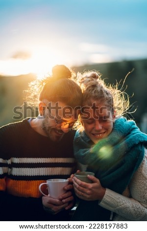Affectionate young couple drinking coffee or tea and hugging while standing on their balcony at home in the morning with a beautiful nature view. Watching the sunrise with a romantic partner.