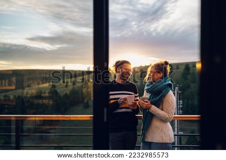 Affectionate young couple drinking coffee or tea while standing on their balcony at home in the morning with a beautiful nature view. Watching the sunrise with a romantic partner. Copy space.