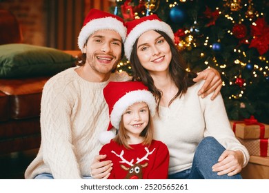 Affectionate young family in Santa hats sitting by Christmas tree together, hugging and smiling at camera - Powered by Shutterstock