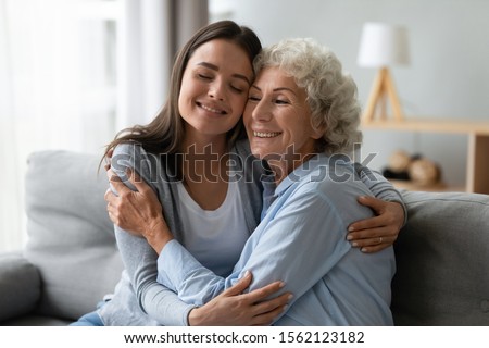 Affectionate young granddaughter and elderly grandma cuddling bonding at home, caring daughter grandchild hugging happy old senior mother enjoying tender moments showing love at home sit on sofa