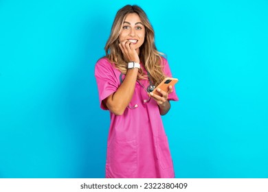 Afraid funny young beautiful doctor woman standing over blue studio background holding telephone and bitting nails