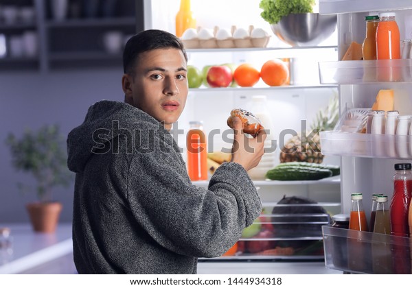 Afraid man caught in the act of eating unhealthy\
food near refrigerator at\
night