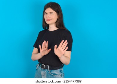 Afraid Young caucasian woman wearing black T-shirt over blue background, makes terrified expression and stop gesture with both hands saying: Stay there. Panic concept. - Shutterstock ID 2154477389