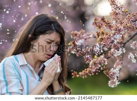 Afraid young woman sneezing in napkin in front of blooming tree. Spring allergy attack concept