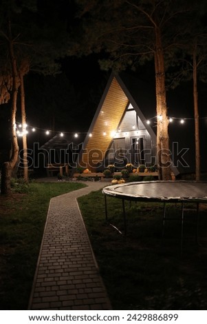 A-frame house in a pine forest. the a-frame house glows with garlands in the evening. rest in the forest. wooden triangle country tiny cabin house in mountains and two chairs.Construction 