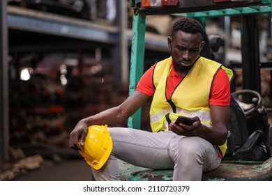 Africa American holding safety helmet and sitting at forklift in the automotive part warehouse. Worker look at smartphone and get the bad news. Feeling stress from fired from work. Disappoint - Powered by Shutterstock