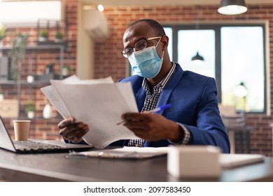 African American Adult Analyzing Documents To Plan Business Project And Strategy. Company Employee Wearing Face Mask, Working With Laptop And Doing Paperwork For Startup During Pandemic.