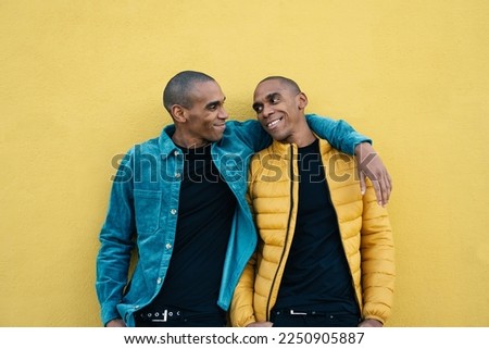 African American adult identical twin brothers hugging and smiling, standing over yellow wall 
