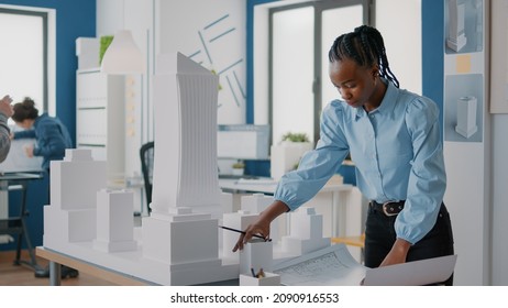 African American Architect Analyzing Blueprints Plan And Building Model At Work. Woman Engineer Working On Construction Layout And Architecture Design To Develop Urban Strategy.