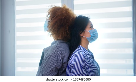 African American And Asian Women With Medical Mask Standing Back To Back In Front Of The Window. Multiracial Friendshp, Mental Health During Covid19. High Quality Photo