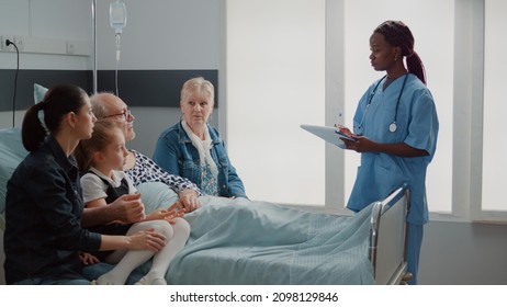 African American Assistant Explaining Medication To Old Man And Visitors In Hospital Ward. Specialist Talking To Family Of Sick Patient About Treatment And Healthcare. Nurse Giving Advice