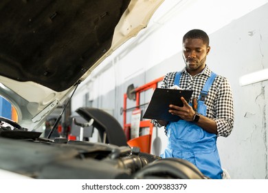 African American auto mechanic man in overalls and holding a notebook in his hands while standing near the open hood of the car