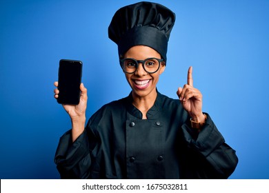 African american baker woman wearing cooker uniform holding smartphone showing screen surprised with an idea or question pointing finger with happy face, number one - Powered by Shutterstock