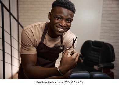 African American barber in working apron is leaning on barber chair - Powered by Shutterstock