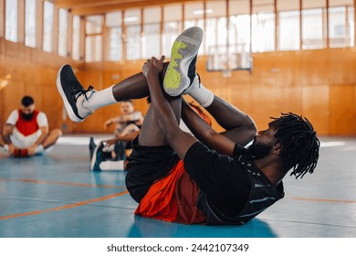 An african american basketball player is lying down on the floor and stretching with his teammates in blurry background during their training. Side view of black man warming up for basketball training - Powered by Shutterstock