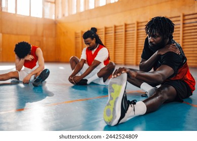 An african american basketball player is sitting on the court and stretching leg. In a blurry background are his multicultural teammates stretching. Interracial basketball team warming up on training. - Powered by Shutterstock