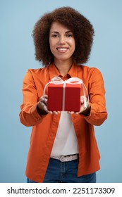 African American beautiful young woman smiles at camera, while holding out a red gift box with a tied white bow, isolated on blue background. A cute present for Saint Valentines Day or any other event - Shutterstock ID 2251173309
