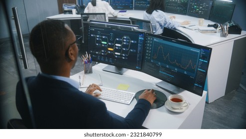 African American big data scientist works at computer in monitoring room. Multiracial colleagues analyze real-time charts at background. Multiple big screens on the wall with displayed server data. - Powered by Shutterstock