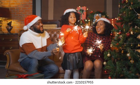 An African American Black Family, Woman, Father, Mother And Kid Girl With Spark Sticks Or Cold Fire, Present Gift In Party On Merry Christmas Eve Xmas On Holiday. Celebration At Home. People Lifestyle