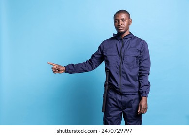 African american bodyguard pointing to something aside in studio, working on an advertisement against blue background. Male security officer in uniform indicates a left or right direction. - Powered by Shutterstock