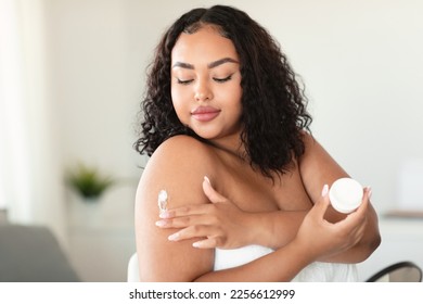 African american bodypositive lady in bath towel applying cream on shoulder, putting on moisturizing body butter at home. Black woman nourishing her skin, enjoying domestic spa procedure - Powered by Shutterstock