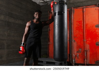 African american boxer with athletic build hugs punching bag wearing red gloves and smiles in dark atmosphere of gym. - Powered by Shutterstock