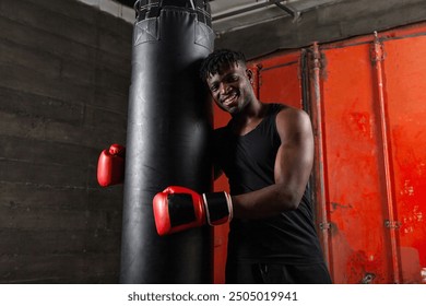 African american boxer with athletic build hugs punching bag wearing red gloves and smiles in dark atmosphere of gym. - Powered by Shutterstock