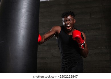 An African American boxer with a furious expression on his face delivers a powerful punch to a punching bag in a dark gym, demonstrating his strength and determination to defeat his opponent in the - Powered by Shutterstock