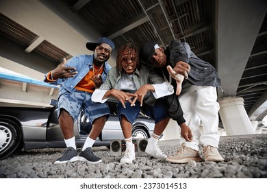 African American boy band consisting of three hip hop performers bending over ground and looking at camera against car with open door - Powered by Shutterstock