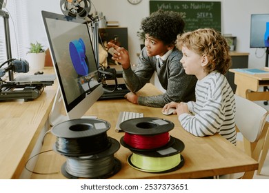 African American boy and Caucasian boy examining 3D printer while sitting in classroom, focusing on creation of objects. 3D printer and filament spools displayed on desk - Powered by Shutterstock