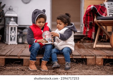 African American boy and girl are sitting at the camper on wooden pallets, playing with a Christmas snow globe. Brother and sister shake the ball, snow flies in it. Christmas holidays outside the city - Powered by Shutterstock