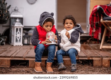 African American boy and girl are sitting at the camper on wooden pallets, playing with a Christmas snow globe. Brother and sister shake the ball, snow flies in it. Christmas holidays outside the city - Powered by Shutterstock