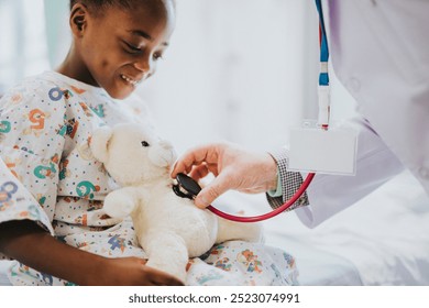 African American boy in a hospital gown smiles, doctor uses a stethoscope on teddy bear. The boy, doctor, and teddy bear create a comforting hospital scene. smiling African American boy in hospital - Powered by Shutterstock