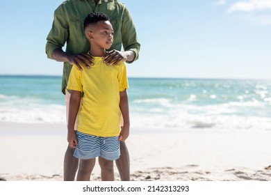 African American Boy Looking Away While Standing Ahead Of Father At Beach Against Sky. Unaltered, Family, Lifestyle, Togetherness, Enjoyment And Holiday Concept.