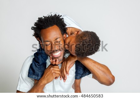 An African American boy on the shoulders of his father, leans over to kiss him on the cheek.  His father laughs with closed eyes.