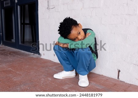 African American boy sits on the ground at school, looking sad. Dressed in casual attire, he rests against a white wall outdoors, conveying a mood of contemplation.