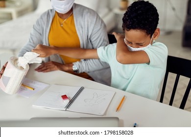 African American boy sneezing into elbow and taking a tissue from a box while learning with mother at home during coronavirus epidemic.  - Powered by Shutterstock