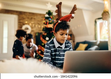 African American Boy Surfing The Net On Laptop While Spending Christmas With His Family At Home.